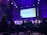 A conference or panel discussion is taking place in a dimly lit auditorium. The audience is seated and watching a speaker panel on stage. A large screen behind the panel displays a presentation titled 'How to Run Successful Advertising for Crypto and Blockchain Projects'. The room is illuminated with purple and blue lighting.