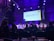 A conference or panel discussion is taking place in a dimly lit auditorium. The audience is seated and watching a speaker panel on stage. A large screen behind the panel displays a presentation titled 'How to Run Successful Advertising for Crypto and Blockchain Projects'. The room is illuminated with purple and blue lighting.