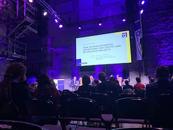 A conference or panel discussion is taking place in a dimly lit auditorium. The audience is seated and watching a speaker panel on stage. A large screen behind the panel displays a presentation titled 'How to Run Successful Advertising for Crypto and Blockchain Projects'. The room is illuminated with purple and blue lighting.