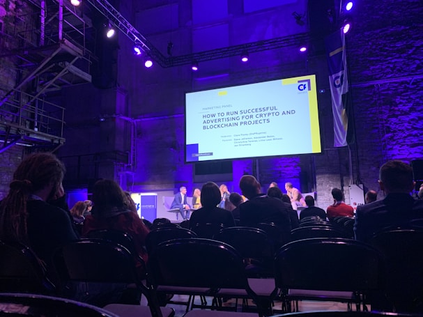 A conference or panel discussion is taking place in a dimly lit auditorium. The audience is seated and watching a speaker panel on stage. A large screen behind the panel displays a presentation titled 'How to Run Successful Advertising for Crypto and Blockchain Projects'. The room is illuminated with purple and blue lighting.
