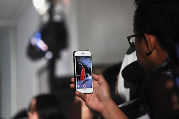 A person is holding a smartphone recording or taking a photo of a fashion show. The phone's screen displays a model walking down a runway wearing a red outfit. The setting appears crowded, suggesting a lively event.