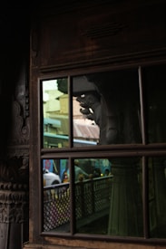 The photograph captures the reflection in an old, intricately carved window. A partial view of a street can be seen through the glass, with a bus and some people in motion. The window's carved frame adds an element of traditional artistry.