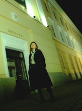 A person with long hair is standing in front of a building at nighttime. The building has light yellow walls and features ornate white decorations above the entrance. The person is wearing a black coat and red lipstick, and they are looking slightly upwards. The building is illuminated with artificial lighting, giving the scene a somewhat dramatic effect.