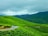 Lush green hills covered with neatly arranged tea plantations under a cloudy sky. A single road winds through the hilly landscape, with a white car traveling along it, possibly carrying bags on the roof. The scene conveys a serene and refreshing rural environment.