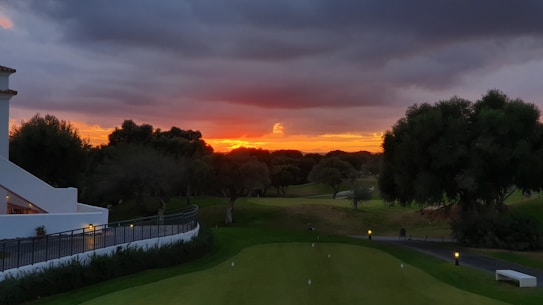 The image depicts a serene landscape during sunset. The sky is a blend of dark clouds with a vivid orange and red horizon. There is a well-maintained green lawn in the foreground, flanked by neatly trimmed bushes and a white building on the left side. Trees are scattered throughout the scene, with the sun casting a warm glow.