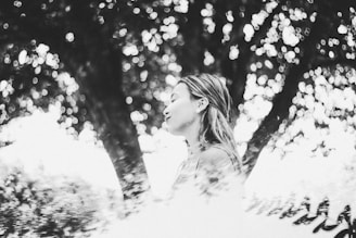 A black and white image featuring a woman in profile with closed eyes amidst a dreamy bokeh effect created by sunlight filtering through tree leaves. The soft focus and light patterns add a poetic and serene quality.