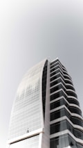A modern high-rise building with curved architecture, featuring a sleek combination of glass and concrete. The structure has multiple floors with dark-tinted windows, creating a striking contrast with the light-colored framework.