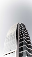 A modern high-rise building with curved architecture, featuring a sleek combination of glass and concrete. The structure has multiple floors with dark-tinted windows, creating a striking contrast with the light-colored framework.