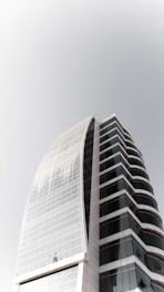 A modern high-rise building with curved architecture, featuring a sleek combination of glass and concrete. The structure has multiple floors with dark-tinted windows, creating a striking contrast with the light-colored framework.