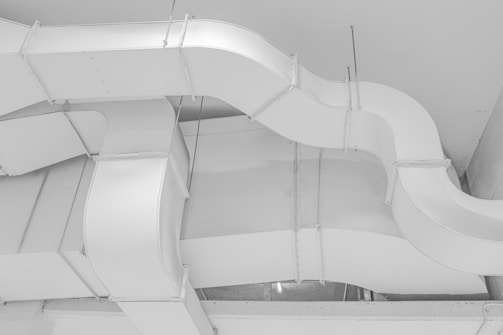 A network of industrial ventilation ducts with a clean, white surface. The ducts are arranged in a curving pattern, highlighting their geometric form. Metal fasteners and joints are visible along the edges, contributing to a sleek, industrial aesthetic.