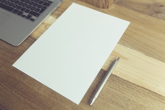 A wooden desk holds an open laptop, a blank sheet of paper, and a silver pen. The composition suggests a workspace or a setting for writing.
