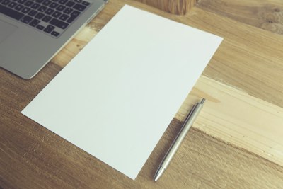A wooden desk holds an open laptop, a blank sheet of paper, and a silver pen. The composition suggests a workspace or a setting for writing.