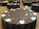 A large banquet hall features multiple round tables covered with black tablecloths. Each table is meticulously set with white plates, silver cutlery, and two salt and pepper shakers, along with numbered signs indicating table numbers. The room is spacious and empty, suggesting preparation for an event.