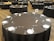 A large banquet hall features multiple round tables covered with black tablecloths. Each table is meticulously set with white plates, silver cutlery, and two salt and pepper shakers, along with numbered signs indicating table numbers. The room is spacious and empty, suggesting preparation for an event.