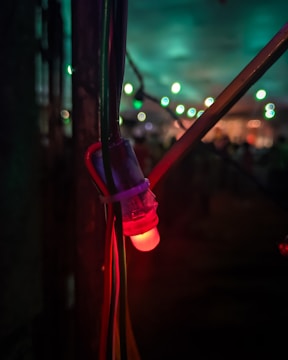 A close-up of a glowing red LED light attached to several cables, set against a dimly lit background with blurred vibrant green and blue lights in the distance.