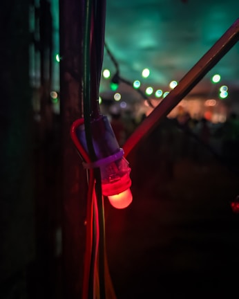 A close-up of a glowing red LED light attached to several cables, set against a dimly lit background with blurred vibrant green and blue lights in the distance.