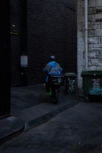 A delivery person wearing a blue jacket and helmet rides a scooter down a dimly lit alley. There are graffiti-covered dumpsters on the side, and the walls are made of old stone and dark brick.