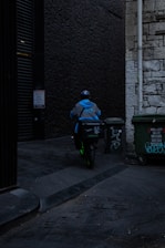 A delivery person wearing a blue jacket and helmet rides a scooter down a dimly lit alley. There are graffiti-covered dumpsters on the side, and the walls are made of old stone and dark brick.
