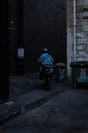 A delivery person wearing a blue jacket and helmet rides a scooter down a dimly lit alley. There are graffiti-covered dumpsters on the side, and the walls are made of old stone and dark brick.