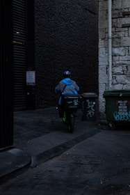 A delivery person wearing a blue jacket and helmet rides a scooter down a dimly lit alley. There are graffiti-covered dumpsters on the side, and the walls are made of old stone and dark brick.