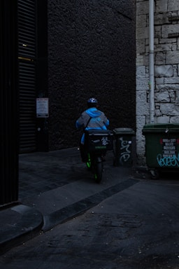 A delivery person wearing a blue jacket and helmet rides a scooter down a dimly lit alley. There are graffiti-covered dumpsters on the side, and the walls are made of old stone and dark brick.