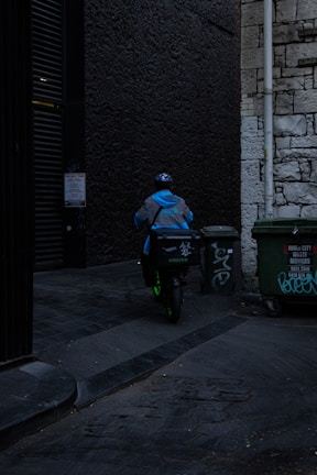 A delivery person wearing a blue jacket and helmet rides a scooter down a dimly lit alley. There are graffiti-covered dumpsters on the side, and the walls are made of old stone and dark brick.