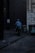 A delivery person wearing a blue jacket and helmet rides a scooter down a dimly lit alley. There are graffiti-covered dumpsters on the side, and the walls are made of old stone and dark brick.
