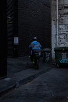 A delivery person wearing a blue jacket and helmet rides a scooter down a dimly lit alley. There are graffiti-covered dumpsters on the side, and the walls are made of old stone and dark brick.