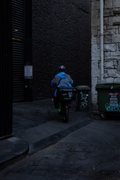 A delivery person wearing a blue jacket and helmet rides a scooter down a dimly lit alley. There are graffiti-covered dumpsters on the side, and the walls are made of old stone and dark brick.