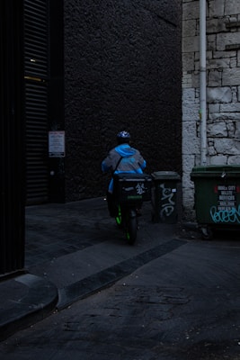 A delivery person wearing a blue jacket and helmet rides a scooter down a dimly lit alley. There are graffiti-covered dumpsters on the side, and the walls are made of old stone and dark brick.