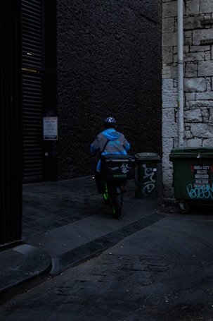 A delivery person wearing a blue jacket and helmet rides a scooter down a dimly lit alley. There are graffiti-covered dumpsters on the side, and the walls are made of old stone and dark brick.