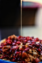 A close-up view of honey being drizzled over a dish consisting of pomegranate seeds and pieces of granola. The honey creates a glossy, smooth effect as it flows downwards. The background is blurred, highlighting the vibrant red of the fruit and the golden tones of the honey.
