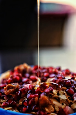 A close-up view of honey being drizzled over a dish consisting of pomegranate seeds and pieces of granola. The honey creates a glossy, smooth effect as it flows downwards. The background is blurred, highlighting the vibrant red of the fruit and the golden tones of the honey.