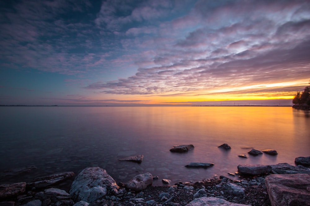 the sun is setting over the ocean with rocks in the foreground