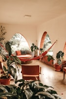 A cozy indoor space with terracotta arched windows and lush green plants. A woven lounge chair with orange and gray cushions is paired with a red hammock. The natural light filters through the windows, casting soft shadows.