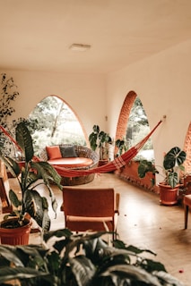A cozy indoor space with terracotta arched windows and lush green plants. A woven lounge chair with orange and gray cushions is paired with a red hammock. The natural light filters through the windows, casting soft shadows.