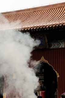 Thick white smoke drifts in front of a traditional building with a tiled roof and ornate, dark-colored door. People are partially visible behind the smoke, one of whom is wearing a brown jacket.