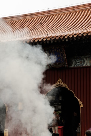 Thick white smoke drifts in front of a traditional building with a tiled roof and ornate, dark-colored door. People are partially visible behind the smoke, one of whom is wearing a brown jacket.