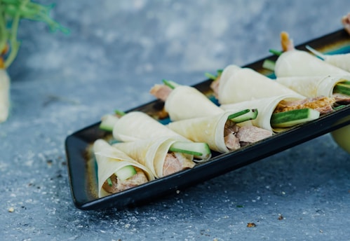 A close-up view of several rolled appetizers on a rectangular black plate. These rolls appear to be made with thinly sliced vegetables wrapping fillings that include slices of cucumber and possibly meat. The background is a textured, mottled blue-gray surface.