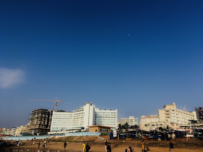 A beachfront with several large buildings in the background, including a partially constructed high-rise featuring a crane. The sky above is clear with a faint moon visible. There are people walking and engaging in activities near the shore.