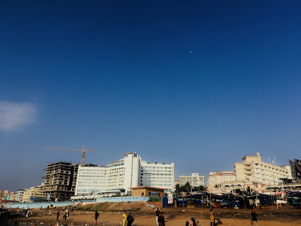 A beachfront with several large buildings in the background, including a partially constructed high-rise featuring a crane. The sky above is clear with a faint moon visible. There are people walking and engaging in activities near the shore.