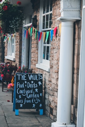 A building exterior with stone walls is adorned with colorful triangular banners. A blackboard sign outside advertises activities like pool and darts, as well as a courtyard garden with beer prices. Hanging flower baskets with vibrant blooms add to the festive atmosphere.