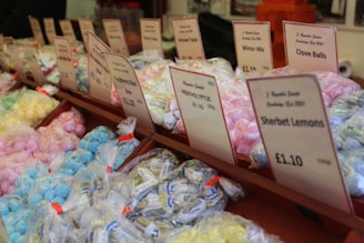 Multiple rows of sweet treats, each packaged in clear plastic bags and neatly organized, are displayed. Labels with product names like 'Sherbet Lemons' and 'Raspberry Bon Bon' along with prices are visible above each section. The sweets are in a variety of pastel colors including blue, pink, yellow, and green.
