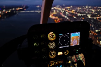 The image captures the inside of a helicopter cockpit at night with illuminated instrument panels displaying various gauges and digital screens. The view through the cockpit window shows a blurred cityscape with lights twinkling, suggesting a city seen from above during flight.