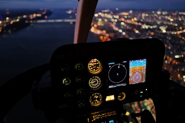 The image captures the inside of a helicopter cockpit at night with illuminated instrument panels displaying various gauges and digital screens. The view through the cockpit window shows a blurred cityscape with lights twinkling, suggesting a city seen from above during flight.