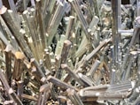 A dense cluster of sharp, elongated metallic crystals with a reflective, shiny surface, appearing in a tightly packed arrangement.