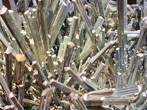 A dense cluster of sharp, elongated metallic crystals with a reflective, shiny surface, appearing in a tightly packed arrangement.
