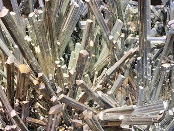 A dense cluster of sharp, elongated metallic crystals with a reflective, shiny surface, appearing in a tightly packed arrangement.