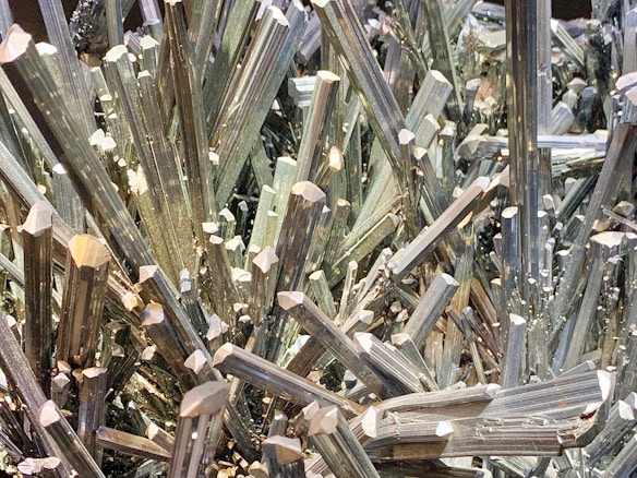 A dense cluster of sharp, elongated metallic crystals with a reflective, shiny surface, appearing in a tightly packed arrangement.