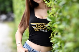 A person with long brown hair is wearing a black tank top with a 'Real Poker' logo in yellow and white, standing partially behind green foliage. They are also wearing blue jeans, showcasing a casual look.